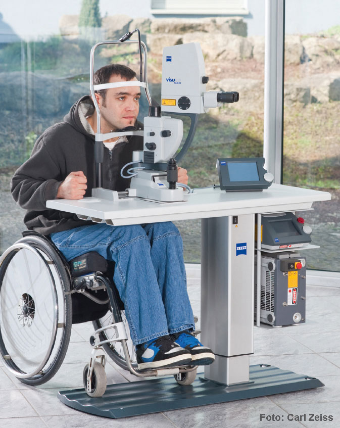 En la imagen se muestra una persona en silla de ruedas en una mesa de examen oftalmológico regulable en altura